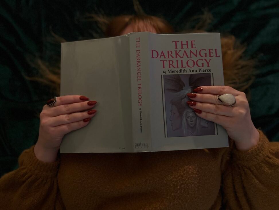A person with red nails and a large ring holds a copy of "The Darkangel Trilogy" by Meredith Ann Pierce, partially covering their face. The setting is cozy and intimate.